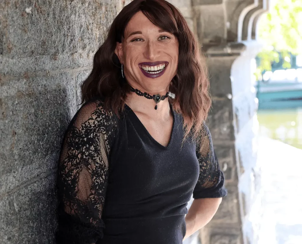 A woman, Nuri, smiles warmly at the camera. She has long, wavy brown hair and is wearing a black top with lace sleeves, a choker necklace, and dark lipstick. She stands against a stone wall with an arched structure in the background, bathed in natural light.