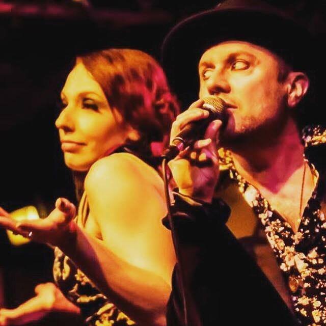 A close-up of two members of the musical group Eddie Japan performing on stage. The man on the right, wearing a black hat and patterned shirt, sings into a microphone, while the woman on the left, with reddish hair, gestures expressively with her hand. The stage lighting casts a warm, golden glow over both performers.