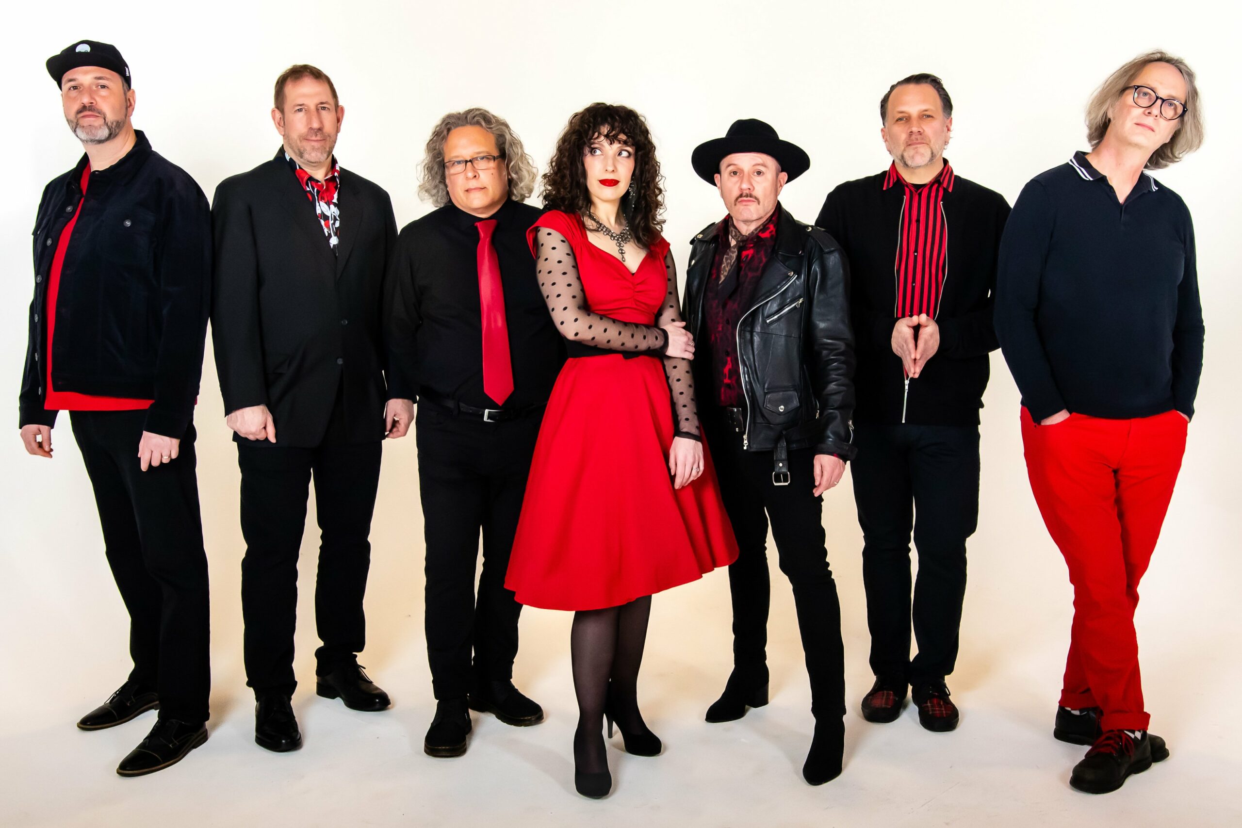 The musical group Eddie Japan poses against a plain background. The group consists of six men and one woman, all dressed in coordinated black and red attire. The woman, standing in the center, wears a red dress with sheer polka dot sleeves, while the men are dressed in a mix of suits, leather jackets, and casual wear, creating a stylish and unified look.