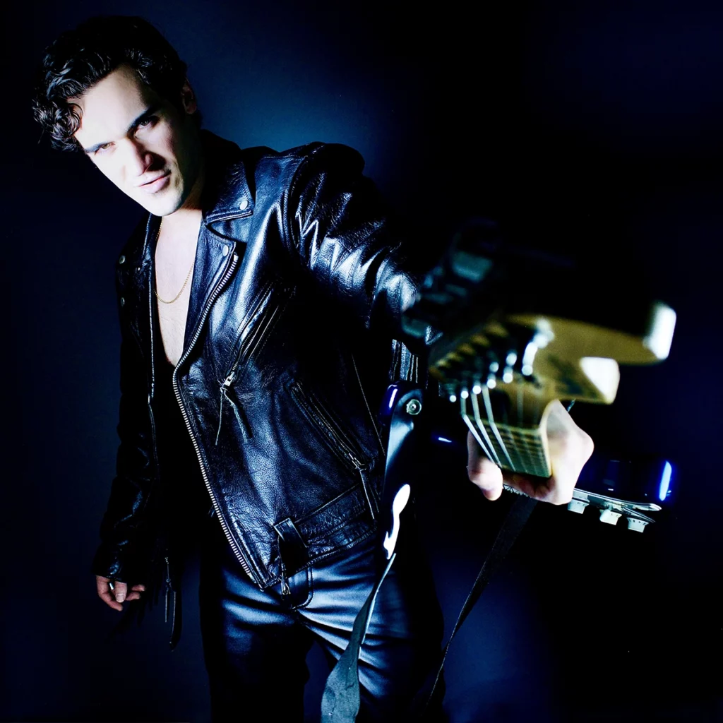 A young man, Jason Damico, dressed in a black leather jacket and leather pants, holds a guitar towards the camera. He has a confident and intense expression, with his dark, wavy hair slightly tousled. The background is dark, making the leather outfit and guitar stand out, creating a bold and edgy atmosphere.