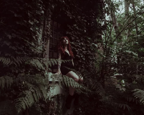 A woman with long red hair, dressed in a black outfit, sits on the windowsill of an ivy-covered building surrounded by dense green foliage. She looks upwards, adding a mysterious and contemplative air to the scene. Ferns and other plants fill the foreground, creating a lush, overgrown atmosphere.