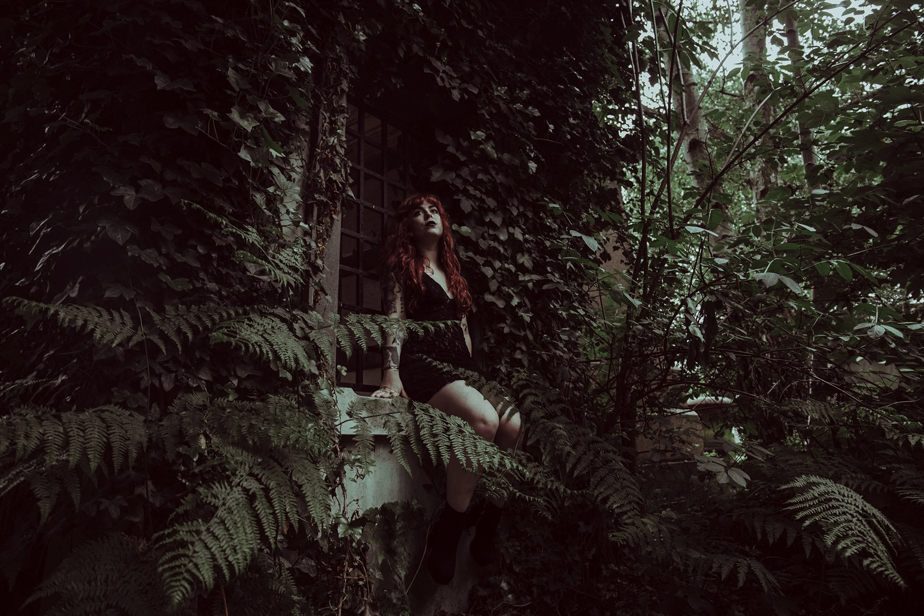 A woman with long red hair, dressed in a black outfit, sits on the windowsill of an ivy-covered building surrounded by dense green foliage. She looks upwards, adding a mysterious and contemplative air to the scene. Ferns and other plants fill the foreground, creating a lush, overgrown atmosphere.