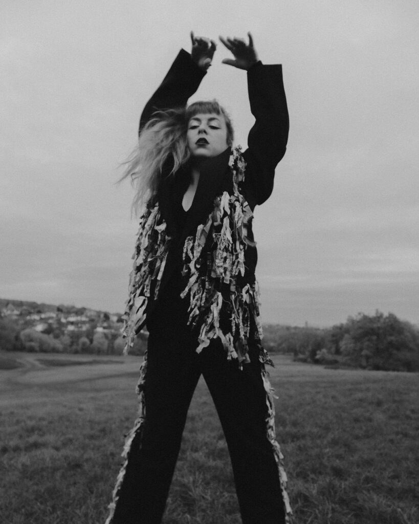 A woman with long, wavy blonde hair stands in a grassy field with her arms raised above her head. She is dressed in a dark outfit adorned with long, hanging fringe. The background is a blurred, rural landscape with trees and hills under a cloudy sky, giving the image a dramatic and moody atmosphere.